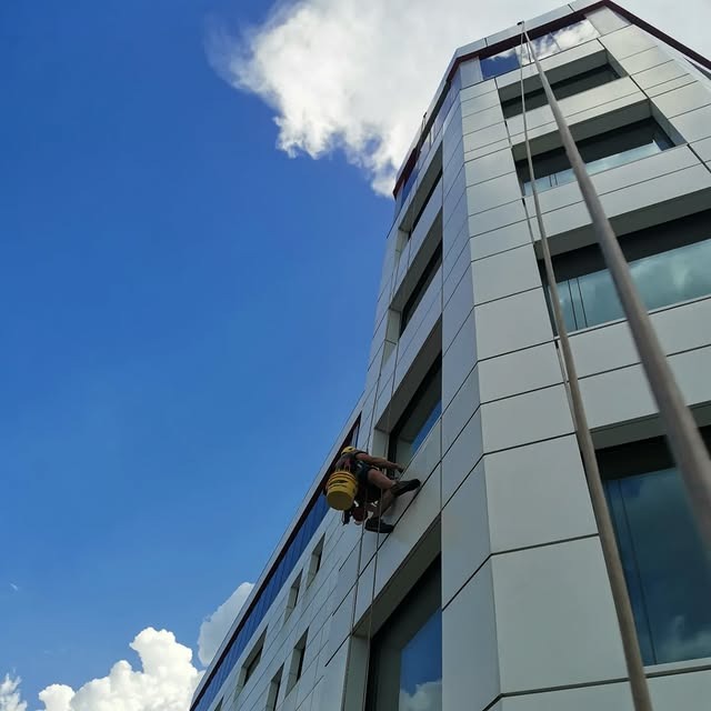 Pracownik na linach, z żółtym wiadrem, myje okna na wysokościowcu o nowoczesnej architekturze, na tle błękitnego nieba z chmurami.