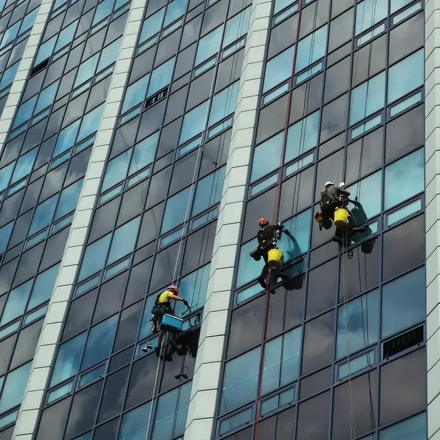 Trzech alpinistów myje okna na wysokościowcu. Pracownicy zawieszeni na linach, z wiadrami i sprzętem do mycia okien. Widok z dołu na szklaną fasadę budynku.