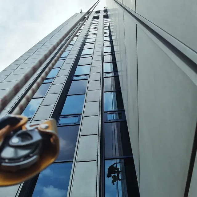 Praca wysokościowa: alpinista w uprzęży, widoczny w odbiciu okien wieżowca, z bliska lina i przyrząd asekuracyjny. Ujęcie z dołu, budynek sięga nieba.