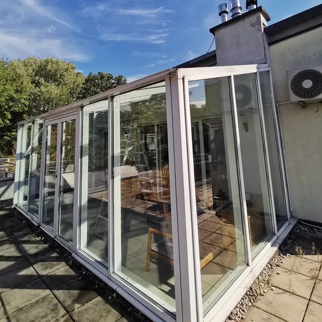 Szklana zabudowa tarasu z widocznym wnętrzem i drabiną, na tle błękitnego nieba. Widoczny klimatyzator na ścianie budynku.