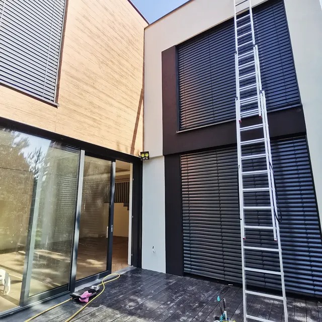 Dwukondygnacyjny dom z ciemnymi żaluzjami i rozłożoną drabiną teleskopową. Widoczne ślady po czyszczeniu na tarasie i oknach. Ujęcie z dołu, w słoneczny dzień.