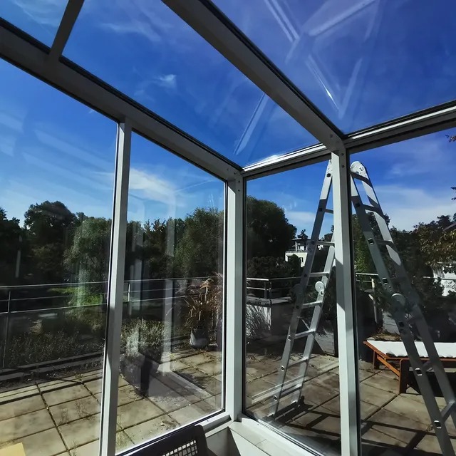 Szklana konstrukcja z widokiem na zewnątrz, drabina oparta o ramę. Błękitne niebo odbija się w czystych szybach.