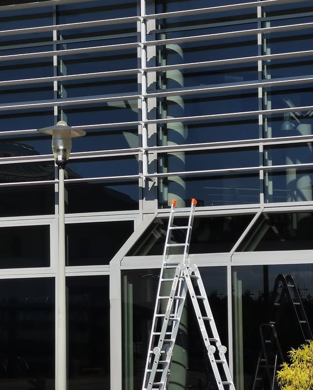 Aluminiowa drabina przystawna oparta o przeszkloną fasadę biurowca z żaluzjami. W tle widoczna latarnia uliczna i fragmenty innych drabin.