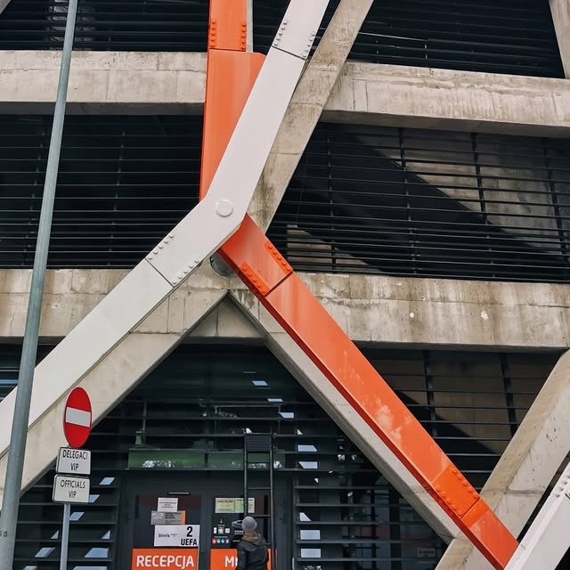 Betonowa fasada budynku z pomarańczowo-białymi elementami konstrukcyjnymi. Widoczne okna z kratami i wejście z napisem 'Recepcja'.