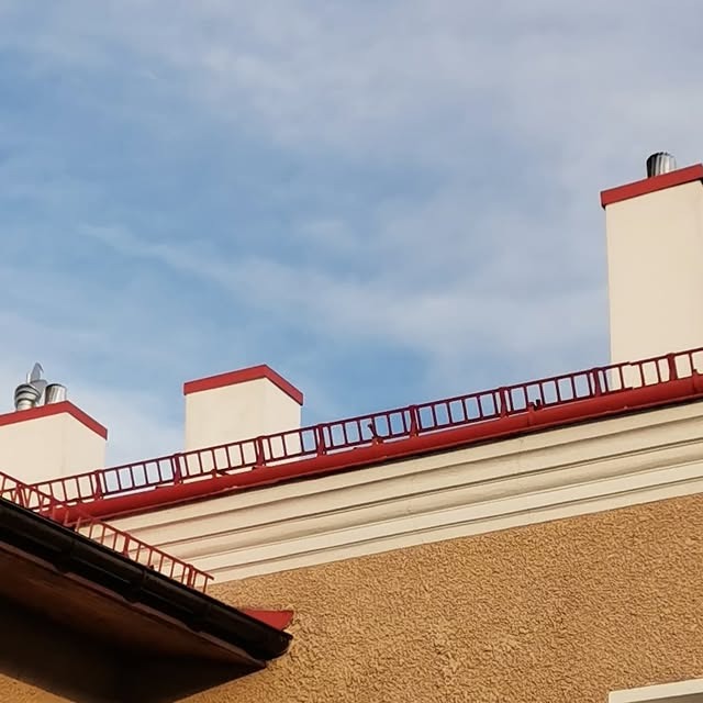 Częściowy widok dachu budynku z kominami i barierkami w kolorze czerwonym na tle błękitnego nieba. Widoczna rynna i elewacja budynku.