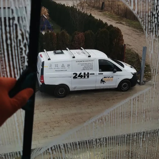 Mycie okna z widokiem na biały samochód dostawczy z logo firmy sprzątającej i napisem 24H, widoczne drzewa w tle, pomarańczowa rękawica trzymająca myjkę.