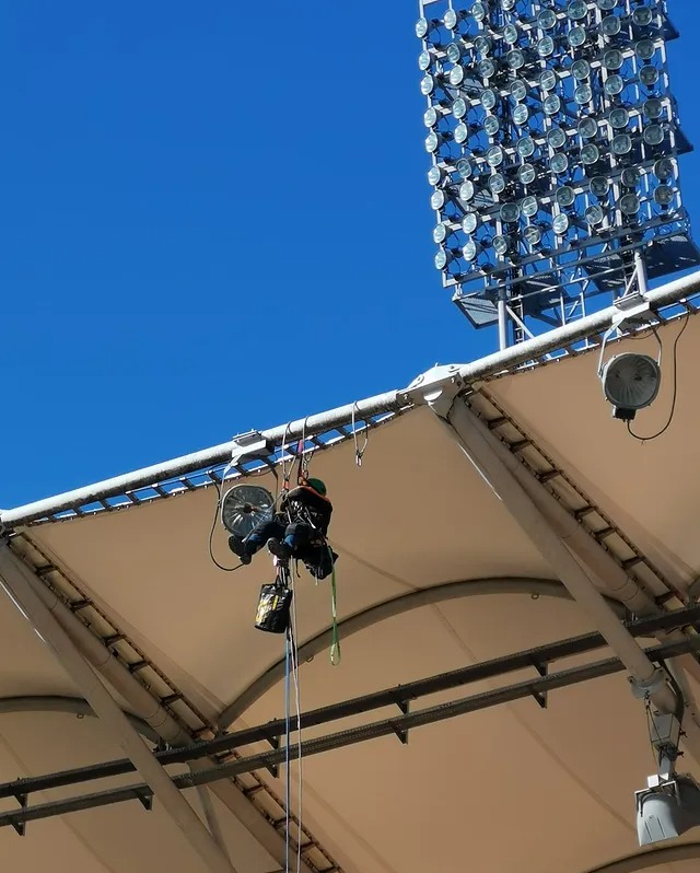 Pracownik na wysokości w Warszawie, zabezpieczony linami, wykonuje prace na konstrukcji dachu stadionu, widoczne oświetlenie. Prace wysokościowe.