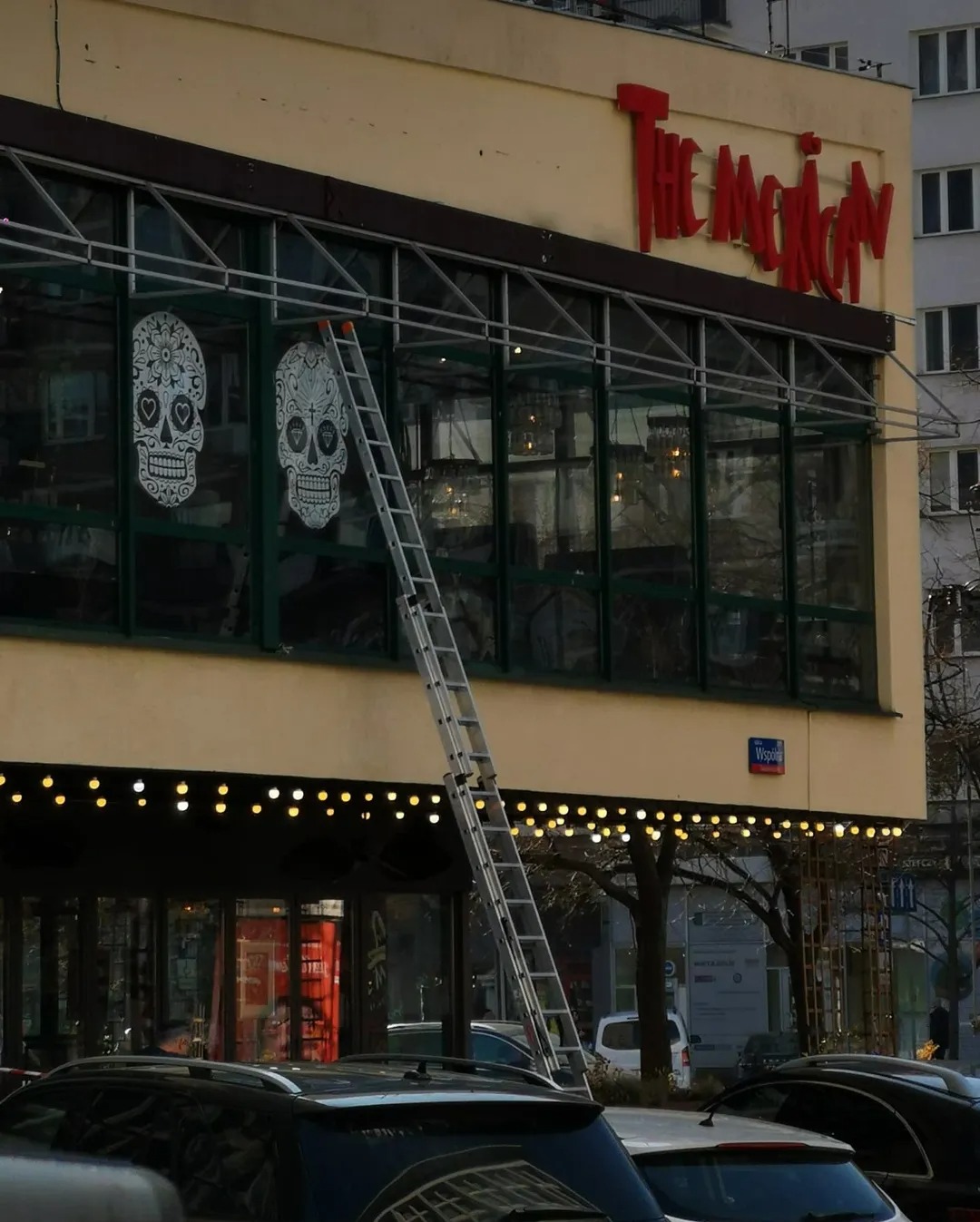 Długa drabina oparta o budynek restauracji 'The Mexican' z dekoracjami czaszek na oknach. Widoczne uliczne oświetlenie i fragmenty zaparkowanych samochodów.