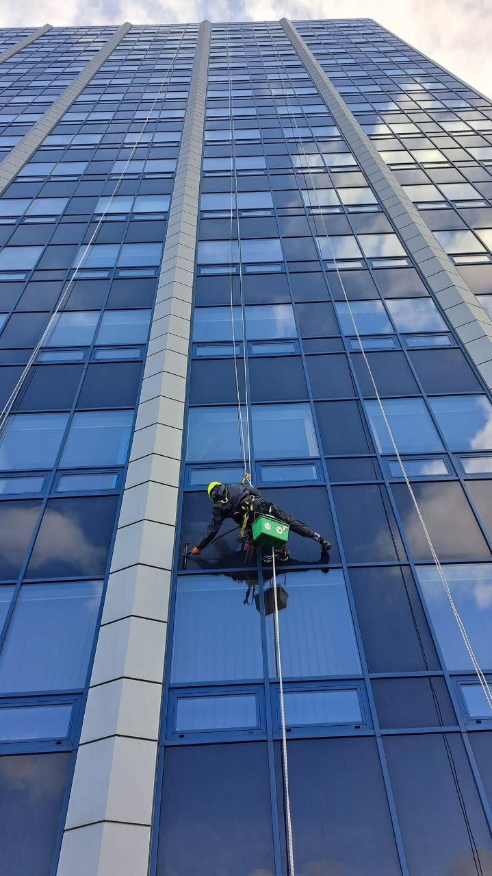 Pracownik w uprzęży, myjący okna wieżowca z niebieskimi ramami, widok z dołu, odbicie chmur w szybach, żółty kask, zielone wiadro na linie.