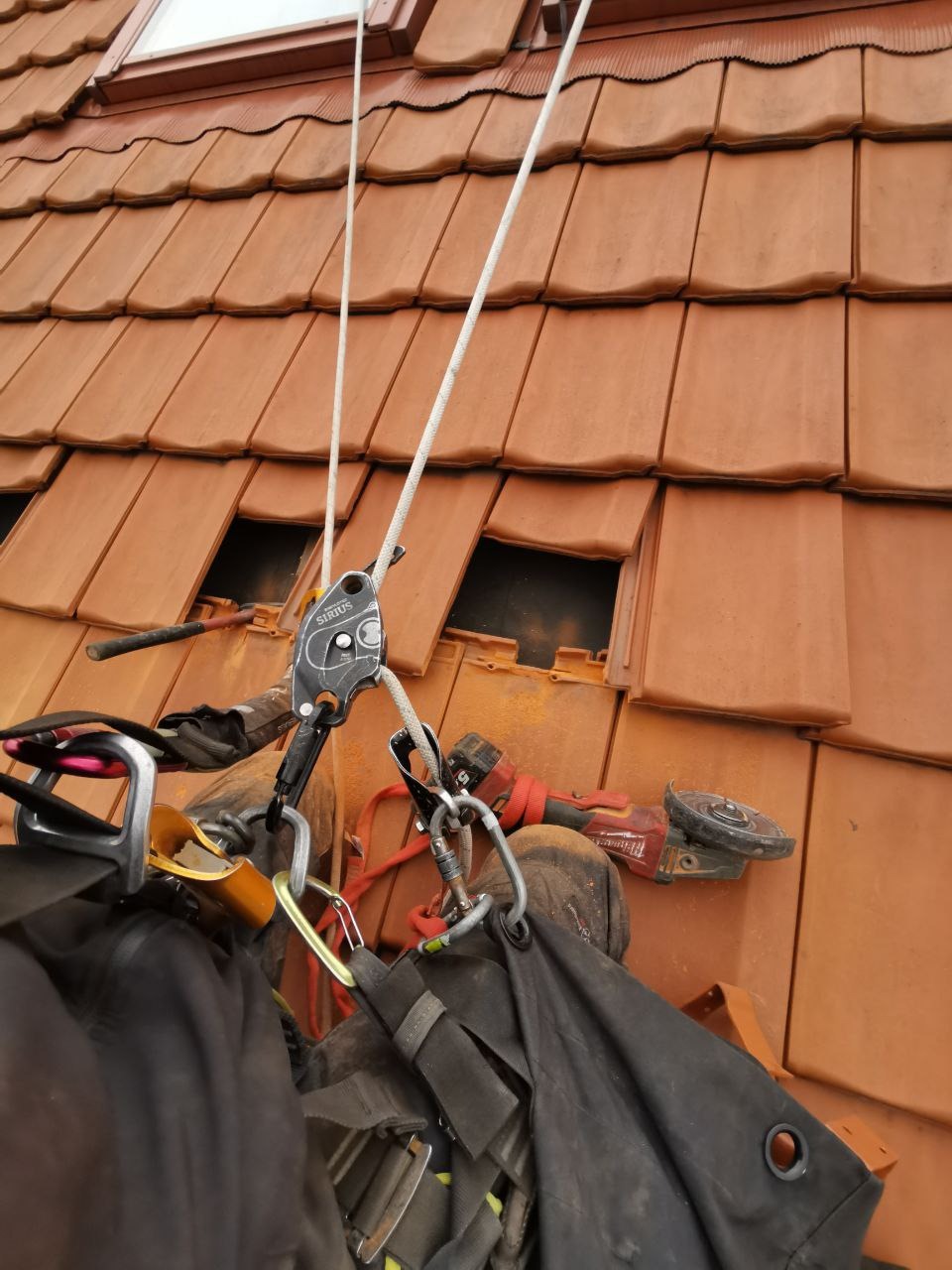 Praca na dachu z użyciem sprzętu alpinistycznego i szlifierki kątowej. Widoczne dachówki, liny, karabinki i narzędzie pracy. Bezpieczeństwo i precyzja podczas naprawy dachu.
