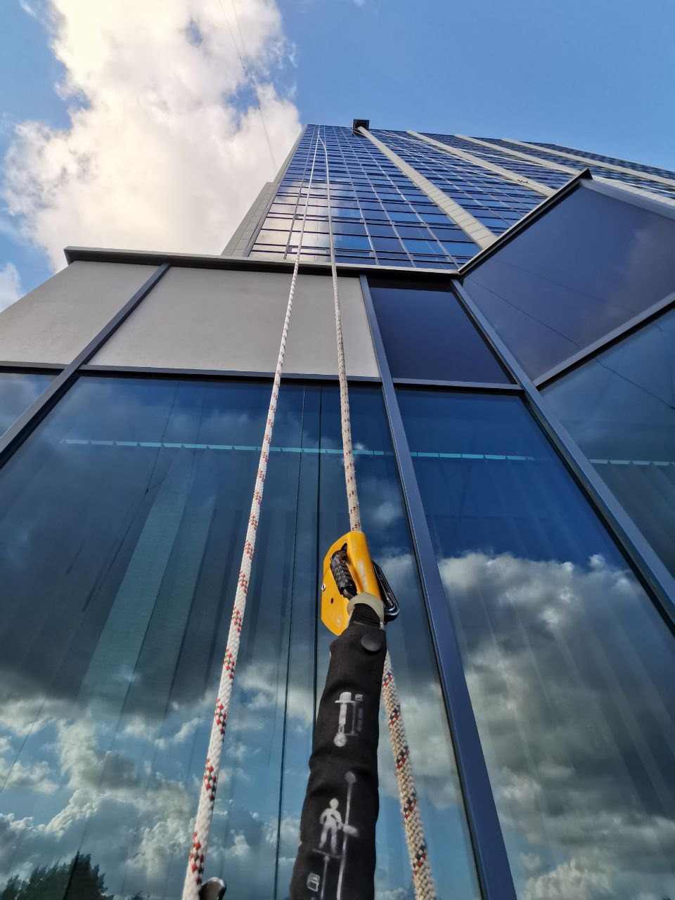 Praca wysokościowa: liny z żółtym przyrządem asekuracyjnym na tle szklanej fasady wieżowca odbijającej błękitne niebo z chmurami. Ujęcie z dołu.
