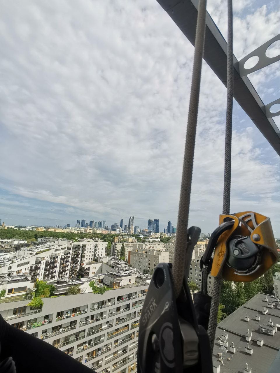 Sprzęt alpinistyczny na tle panoramy miasta z wysokości. Widoczne liny, przyrządy asekuracyjne i budynki w oddali pod błękitnym niebem.