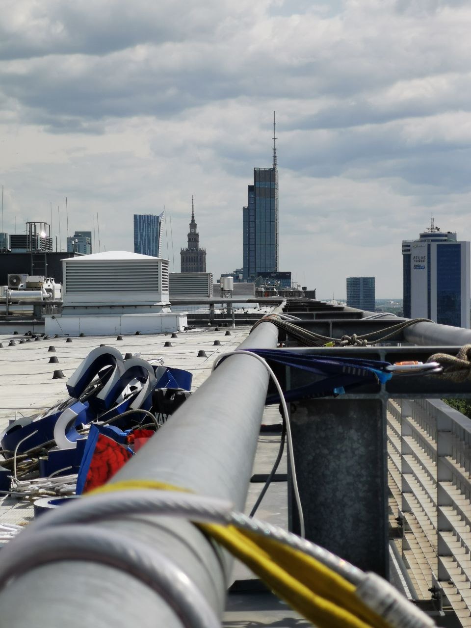 Widok z dachu na panoramę miasta z elementami zabezpieczeń linowych i fragmentami demontowanych liter reklamowych. Perspektywa z wysokości, pochmurne niebo.