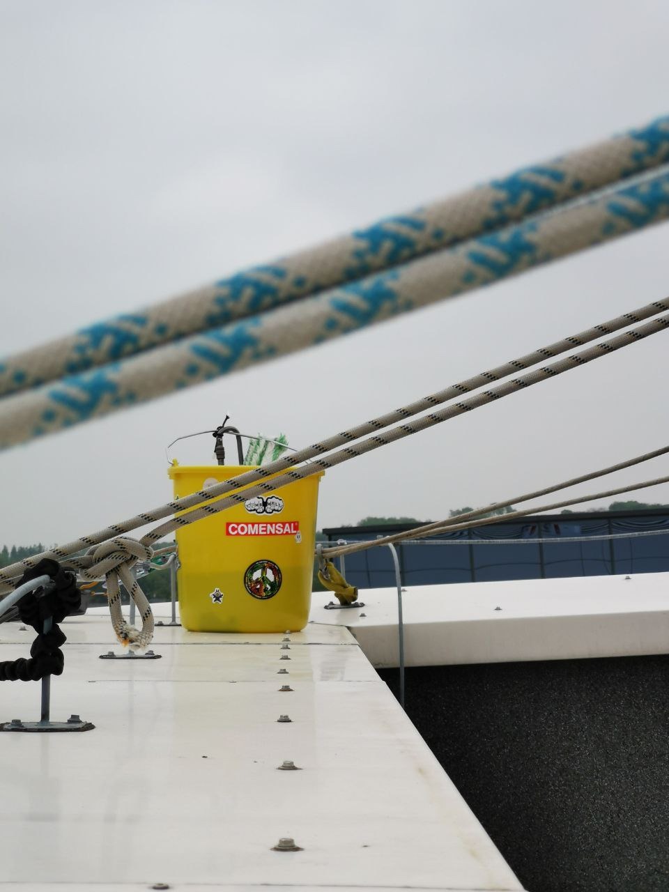 Żółte wiadro z naklejkami na białej powierzchni, liny w tle, sugerujące prace na wysokości. Widok z góry, pochmurne niebo.