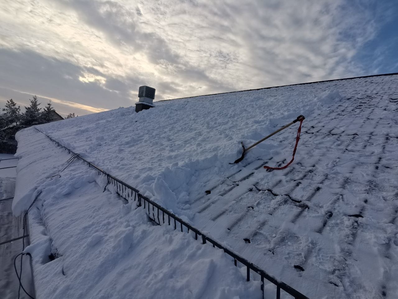 Częściowo odśnieżony dach pokryty śniegiem z widoczną łopatą i barierkami przeciwśniegowymi. W tle pochmurne niebo i komin.