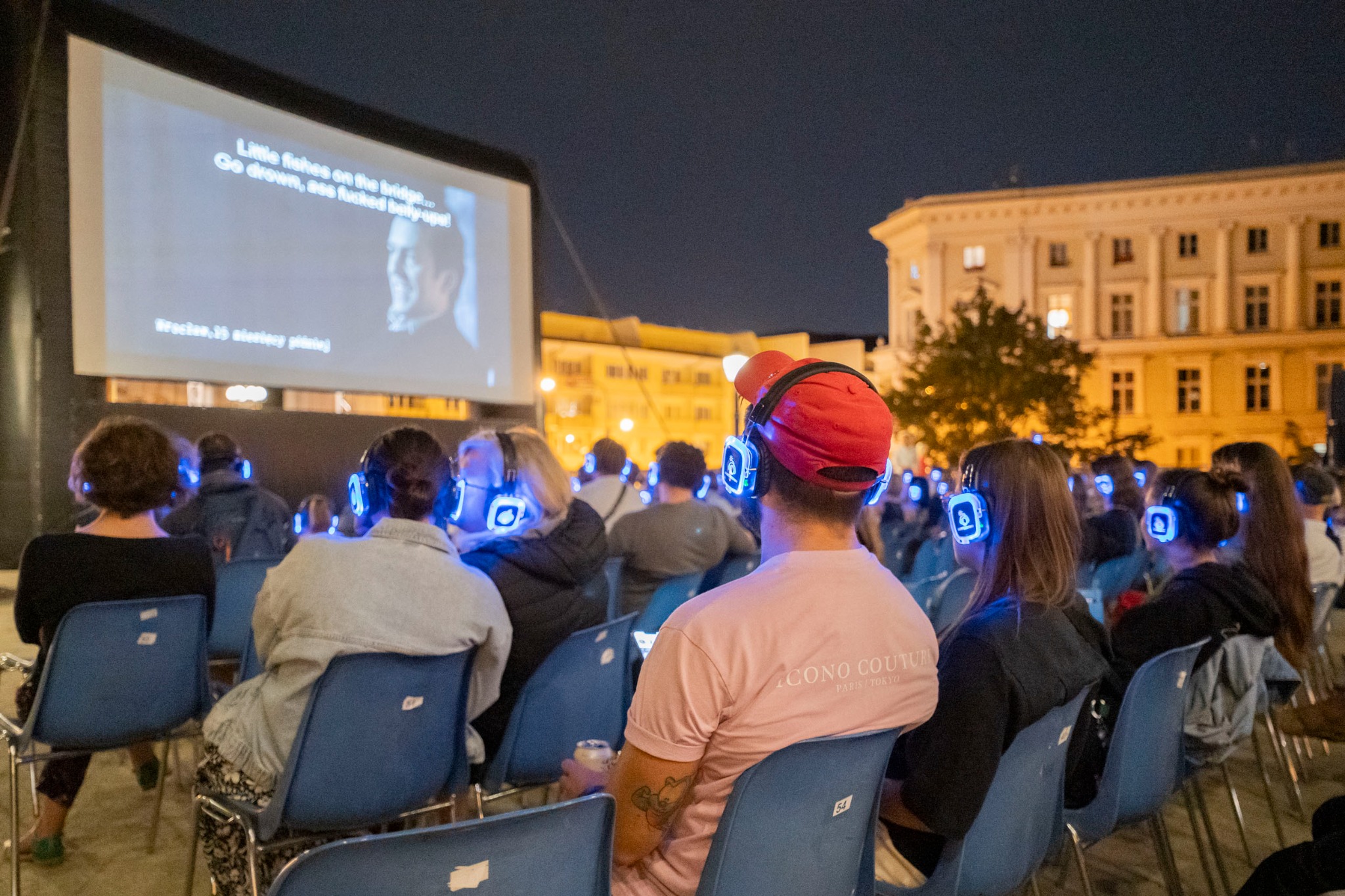 Plenerowe kino nieme z nagłośnieniem słuchawkowym. Widzowie w słuchawkach LED siedzą na krzesłach przed ekranem. W tle budynek.