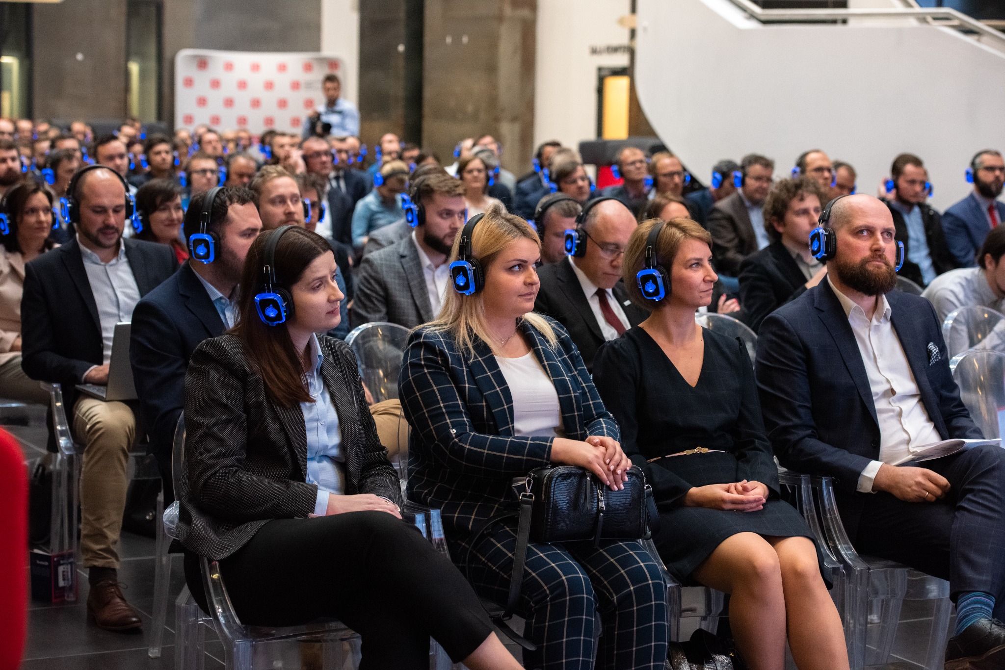 Uczestnicy konferencji w słuchawkach z podświetleniem LED siedzą na przezroczystych krzesłach. W tle widoczny baner i inne osoby.
