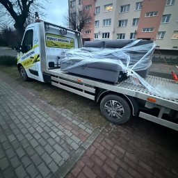 Jakub Piekarski - Laweta z kanapą owiniętą folią na platformie, zaparkowana przy chodniku z kostki brukowej, na tle bloku mieszkalnego. Usługa transportowa mebli.