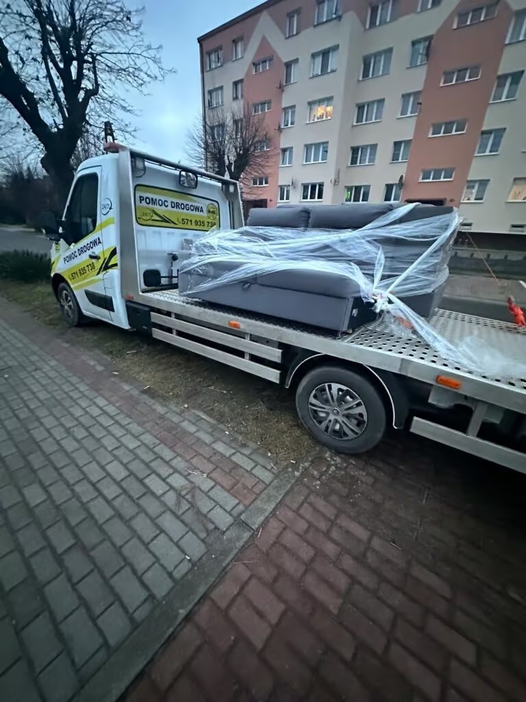 Laweta z kanapą owiniętą folią na platformie, zaparkowana przy chodniku z kostki brukowej, na tle bloku mieszkalnego. Usługa transportowa mebli.