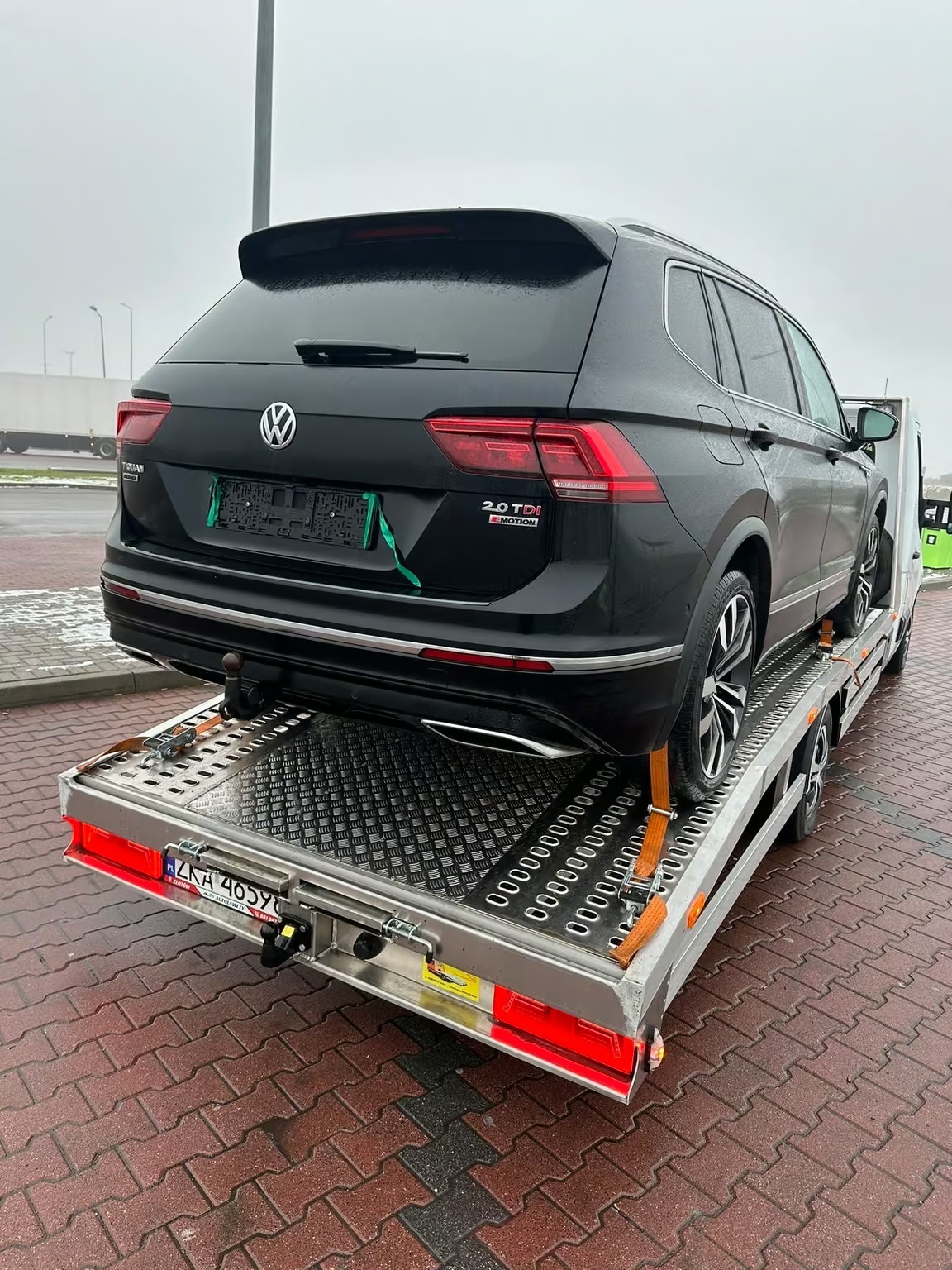 Czarny SUV Volkswagen Tiguan 2.0 TDI eMotion, zabezpieczony pasami, transportowany na srebrnej lawecie z aluminiową platformą. Widok z tyłu, pochmurne niebo w tle.