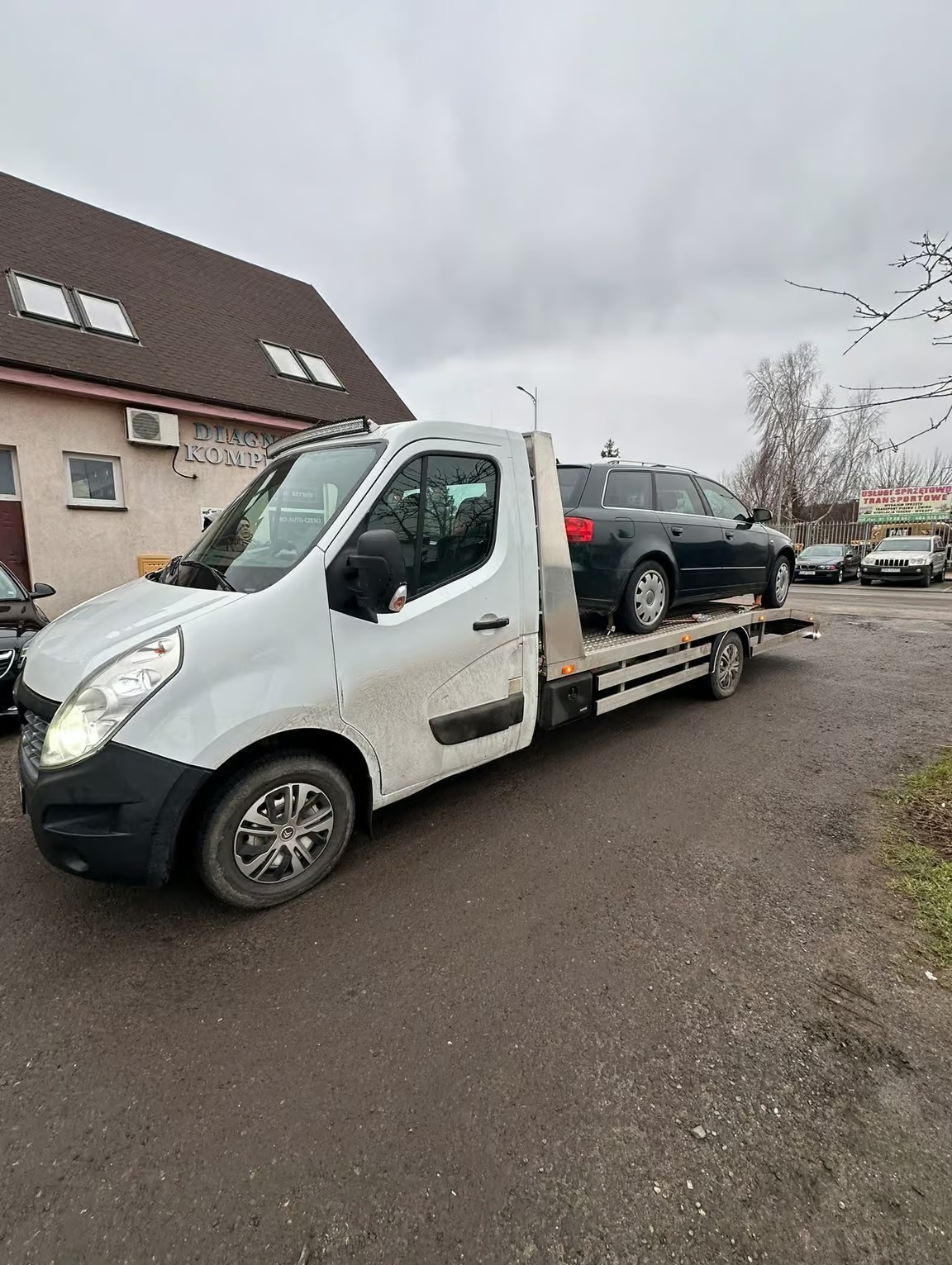Biała laweta Renault Master z załadowanym ciemnym kombi Audi na platformie, zaparkowana na placu przed budynkiem w pochmurny dzień. Widoczny znak 'DIAGNOSTYKA KOMPUTEROWA'.