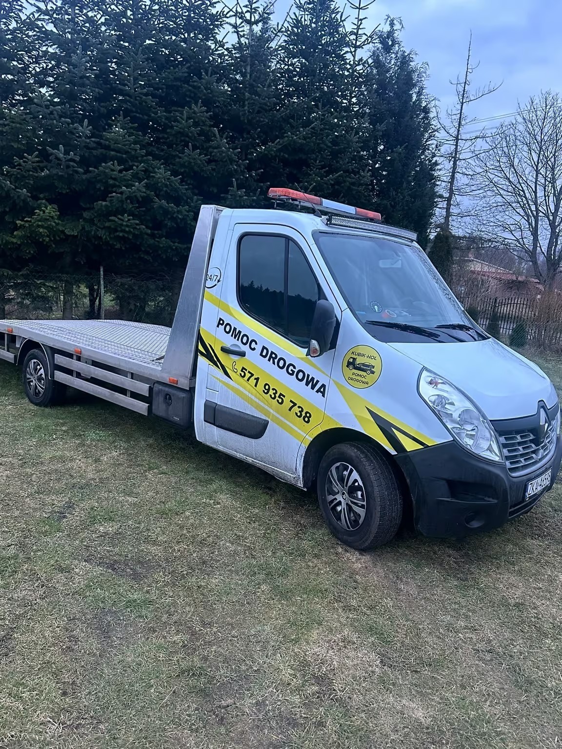 Biała laweta z żółtymi akcentami i napisem 'Pomoc Drogowa' na trawiastym terenie. Widoczne światła ostrzegawcze na dachu i numer telefonu.