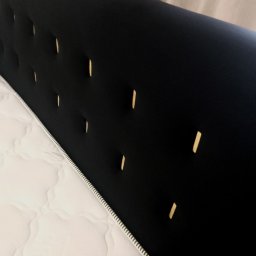 MS Glamour Usługi tapicerskie - Czarne, tapicerowane wezgłowie łóżka z dekoracyjnymi wgłębieniami, widoczne obok białego materaca. Minimalistyczny design i stonowana kolorystyka.