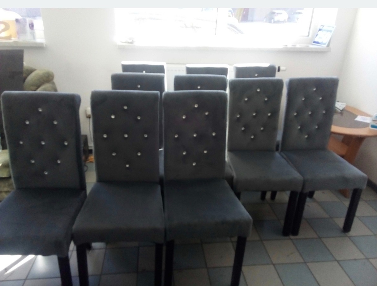 Szereg krzeseł z szarej tkaniny, zdobionych kryształkami, ustawionych w rzędzie na tle jasnego pomieszczenia. Widoczne detale wykończenia i symetryczny układ.