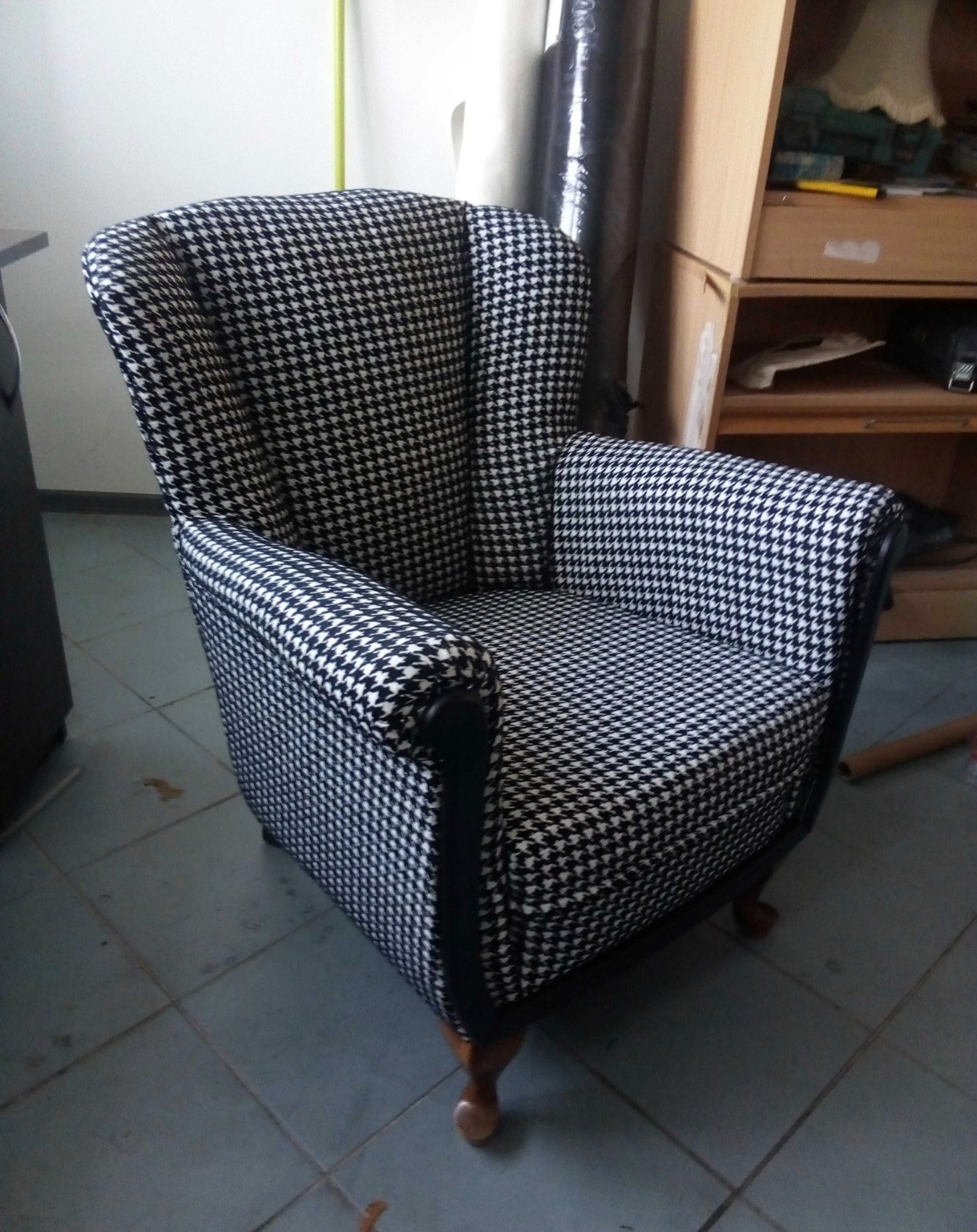 Fotel klubowy po renowacji, obity tkaniną w czarno-białą pepitkę. Widoczne drewniane nóżki i czarne, lakierowane elementy ramy. W tle meble i okno.