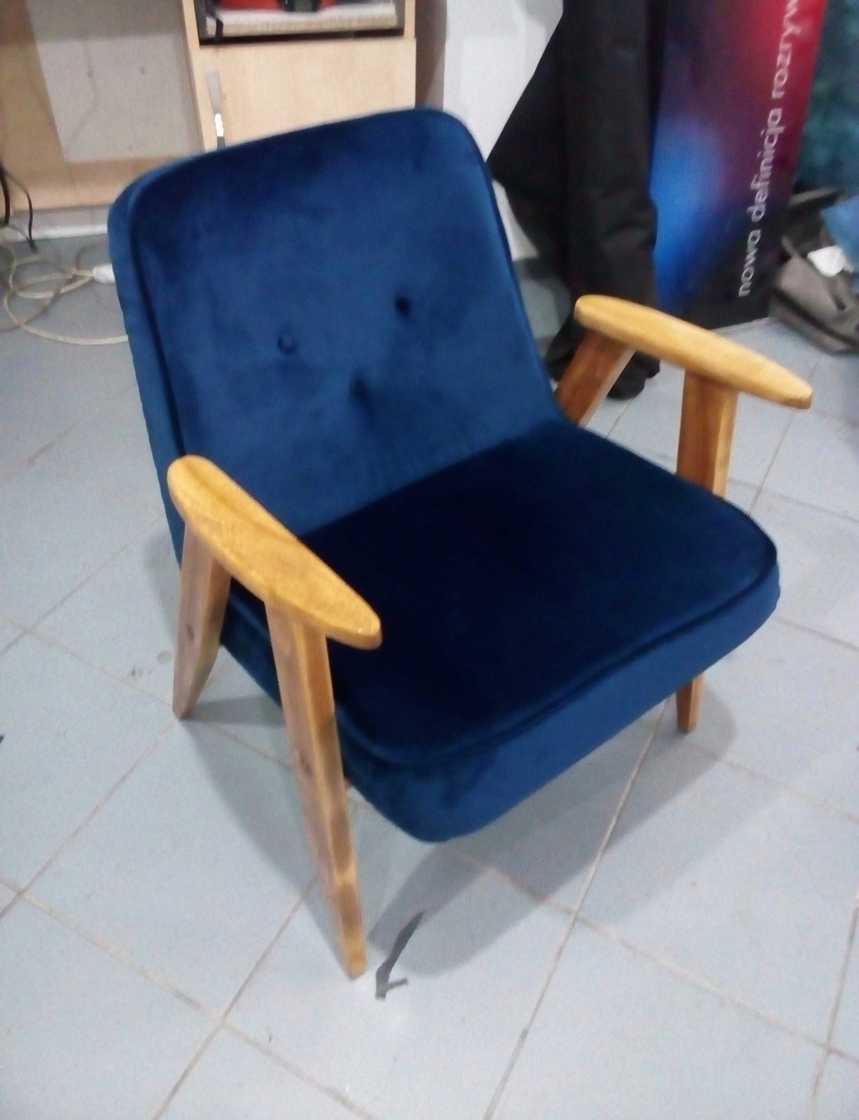 Granatowy fotel w stylu retro z drewnianymi podłokietnikami i nogami, widoczny na tle pomieszczenia z płytkami podłogowymi.
