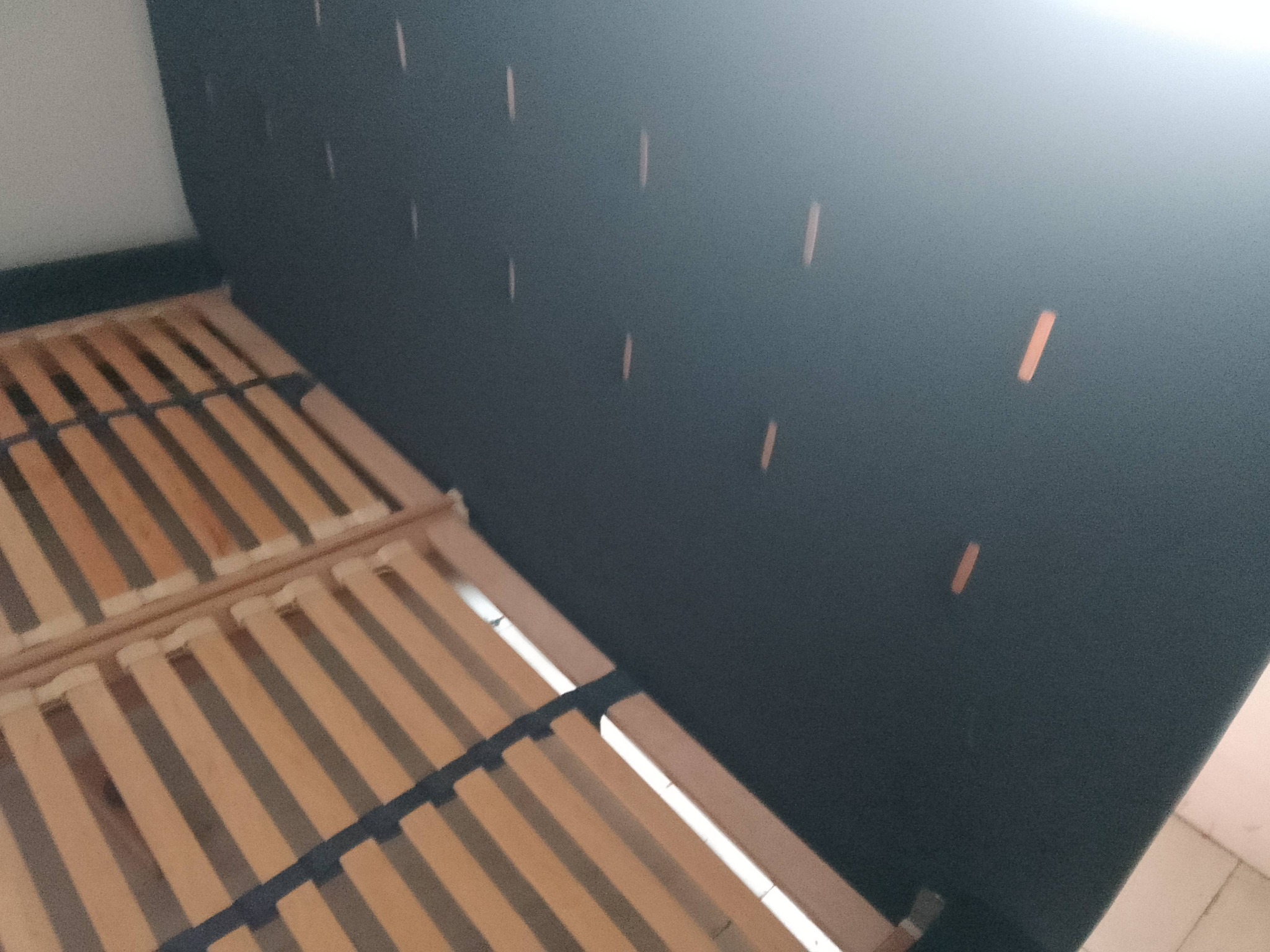 Tapicerowane wezgłowie łóżka w kolorze grafitowym z drewnianym stelażem. Widoczne zszywki na materiale. Ujęcie z bliska, ukazujące detale wykończenia.