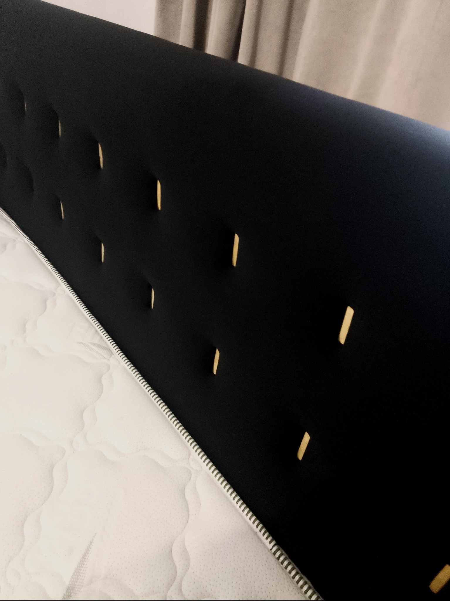 Czarne, tapicerowane wezgłowie łóżka z dekoracyjnymi wgłębieniami, widoczne obok białego materaca. Minimalistyczny design i stonowana kolorystyka.