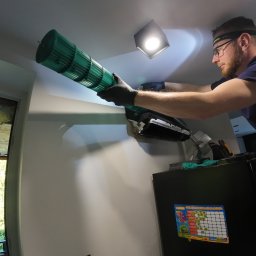 Kalte Luft - Mężczyzna w okularach i rękawicach czyści zielony wentylator klimatyzacji w pomieszczeniu z widocznym oknem i lodówką. Używa latarki czołowej.