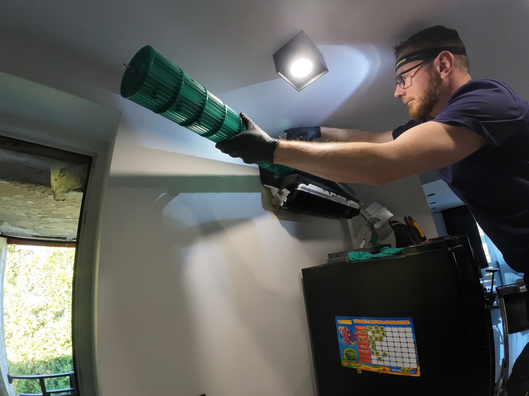 Mężczyzna w okularach i rękawicach czyści zielony wentylator klimatyzacji w pomieszczeniu z widocznym oknem i lodówką. Używa latarki czołowej.