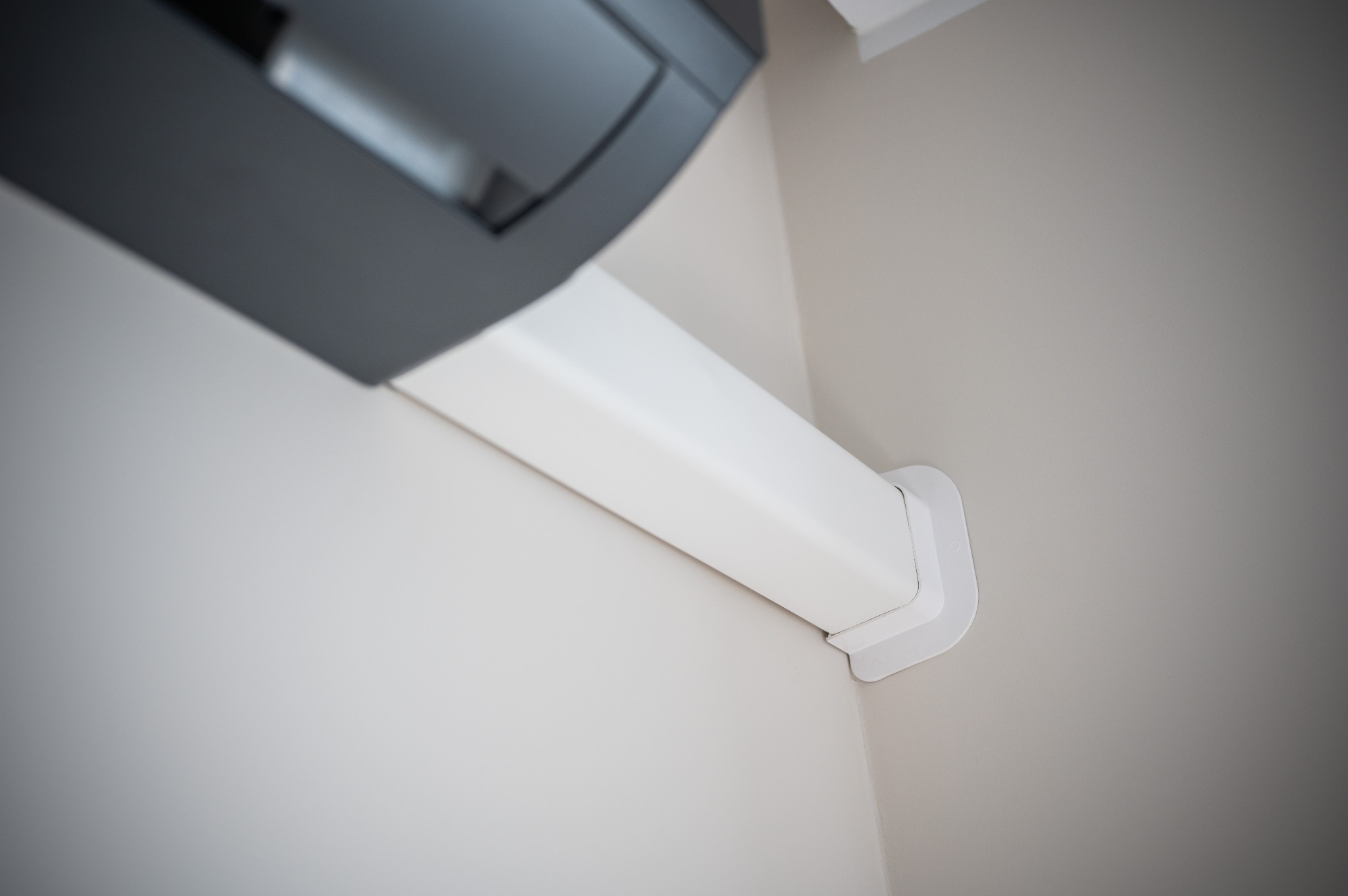 Szary klimatyzator z białą maskownicą rur, zamontowany w rogu jasnego pomieszczenia. Widok z dołu, podkreślający minimalistyczny design i estetyczne wykończenie.