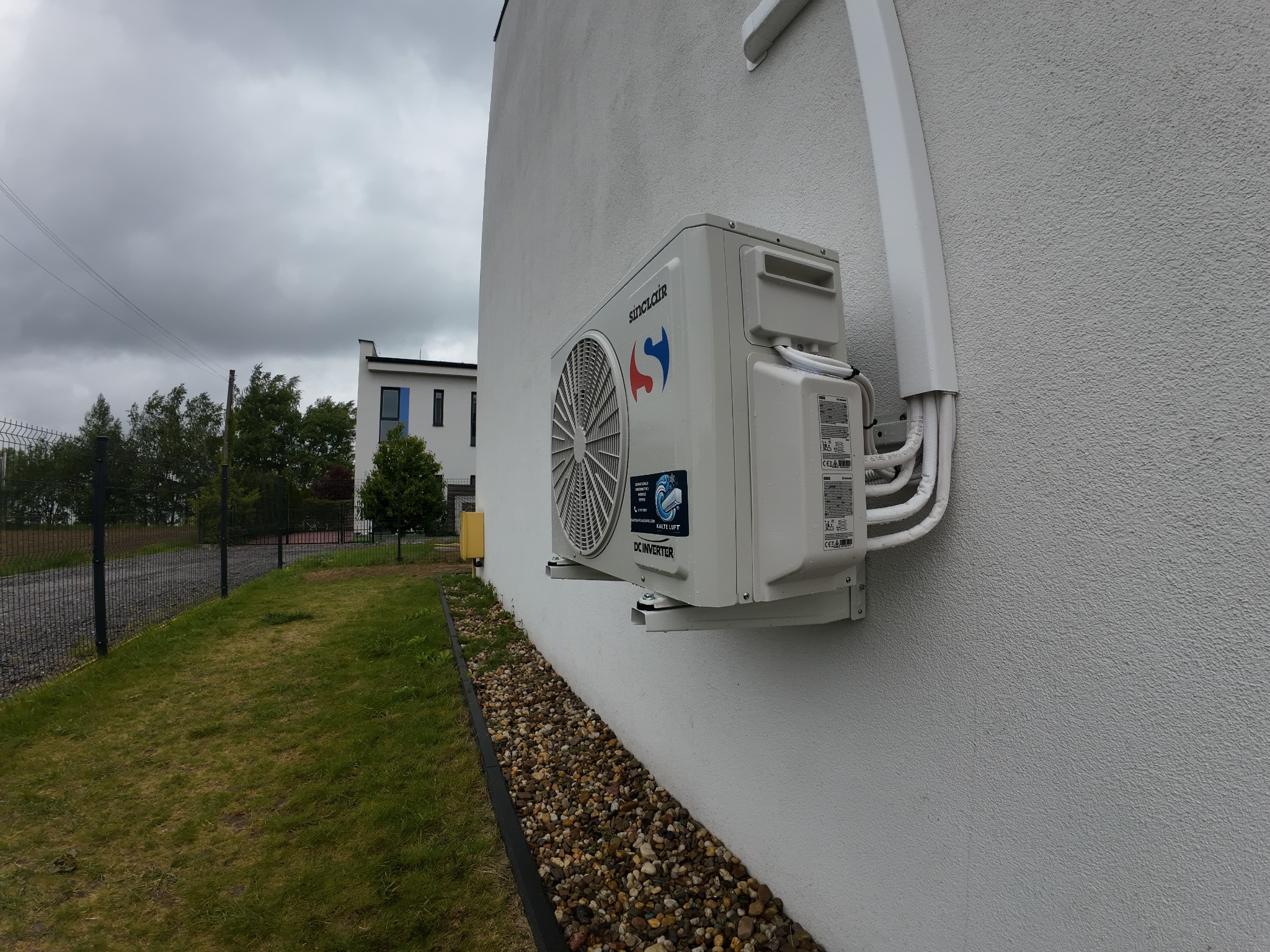 Biała jednostka zewnętrzna klimatyzacji Sinclair zamontowana na elewacji budynku. Widoczne przewody w białej osłonie. Trawa i kamienie dekoracyjne u podstawy ściany.