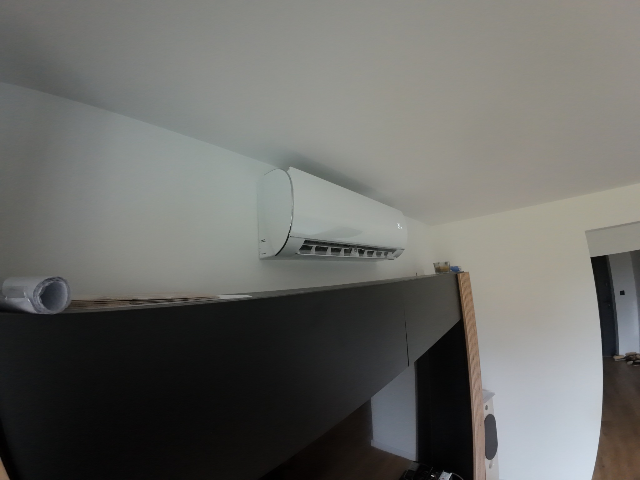 Biały klimatyzator ścienny zamontowany w nowoczesnym wnętrzu, widoczny z perspektywy dolnej, z elementami mebli w tle i fragmentem otwartego przejścia do innego pomieszczenia.