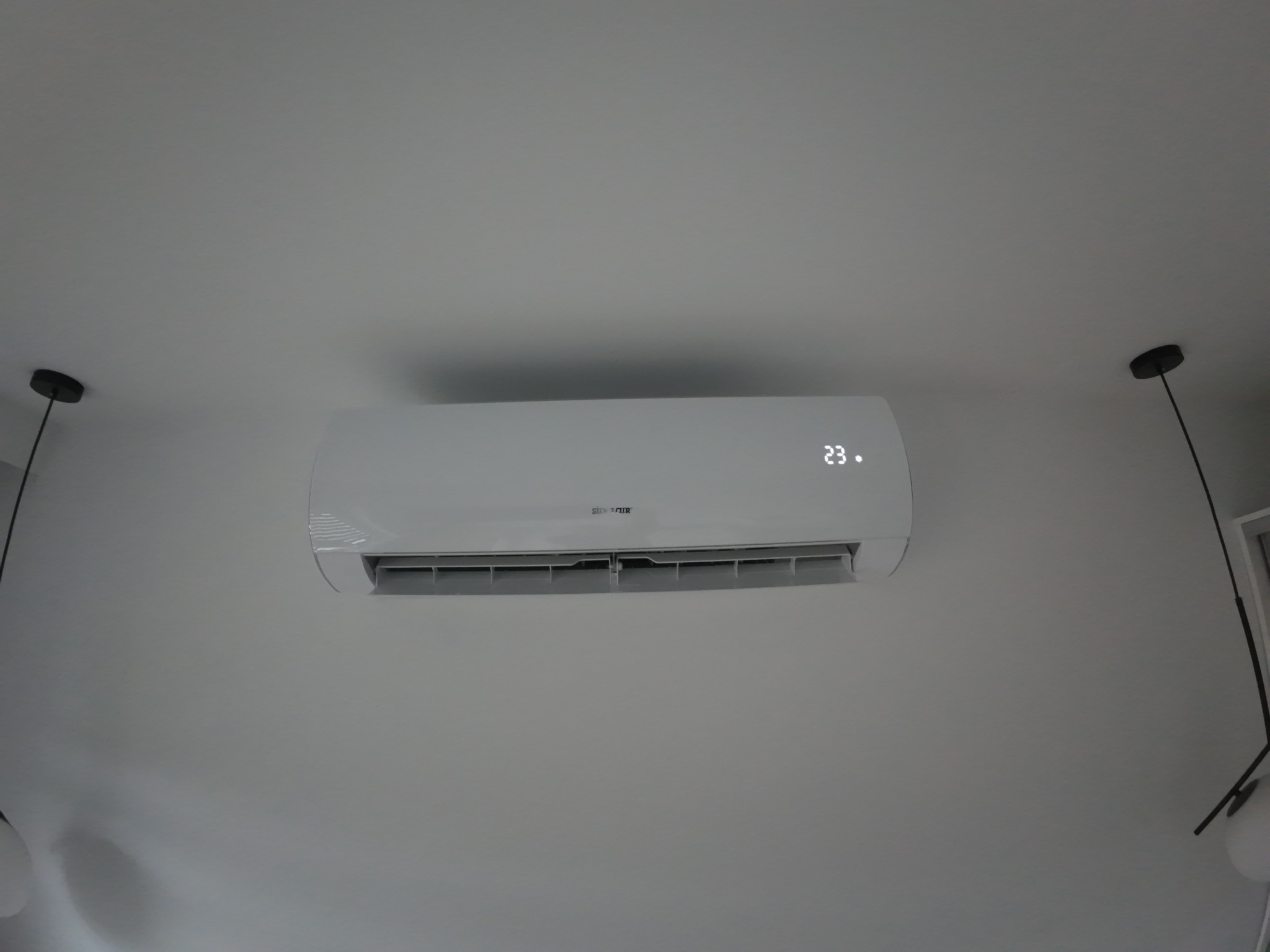 Biały klimatyzator ścienny z wyświetlaczem temperatury 23 stopnie, zamontowany w jasnym pomieszczeniu z lampami po bokach.