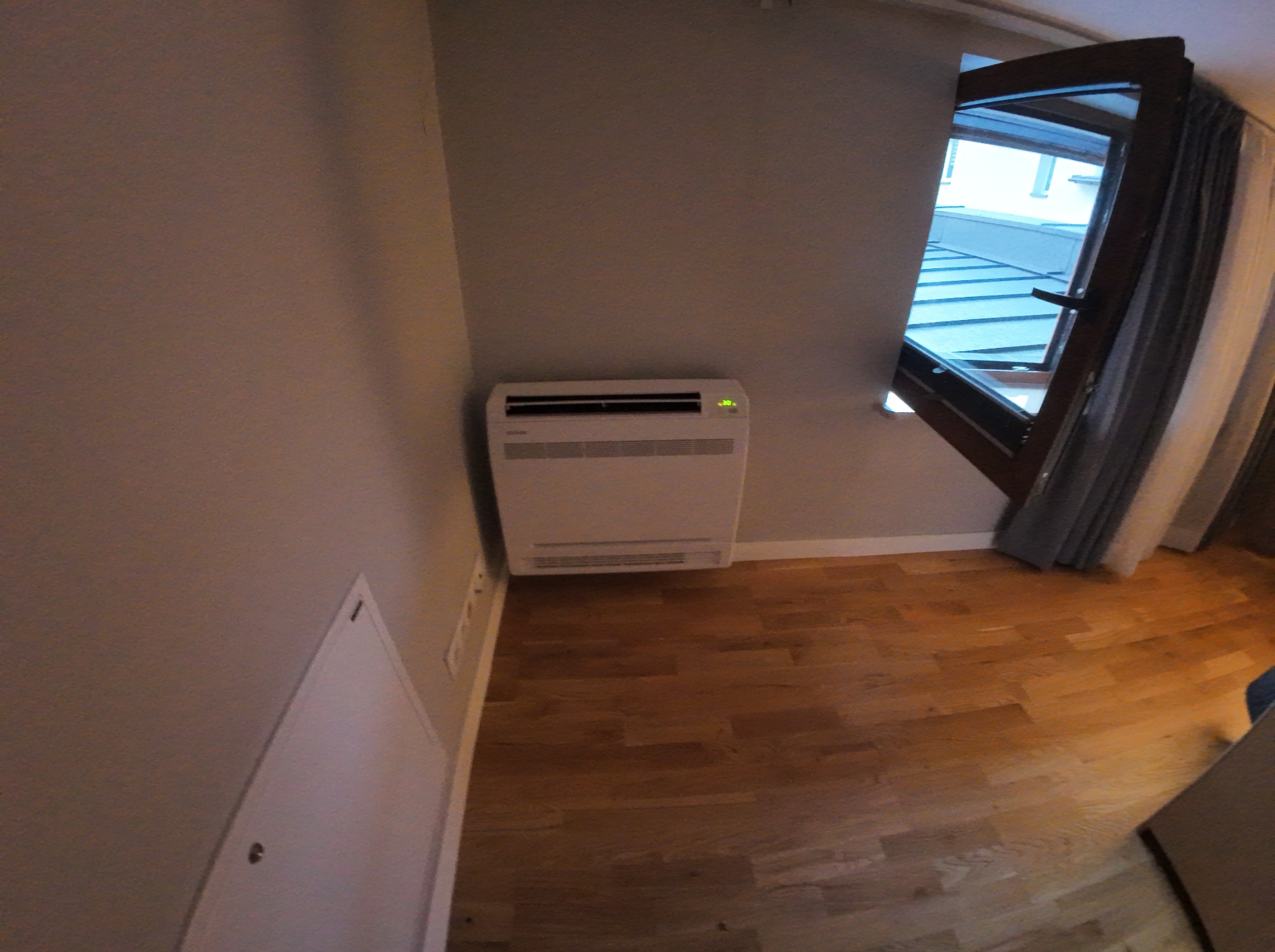 Biały klimatyzator ścienny w jasnym pokoju z drewnianą podłogą, obok okna z zasłoną. Widok z góry, lekko zniekształcony obiektywem.