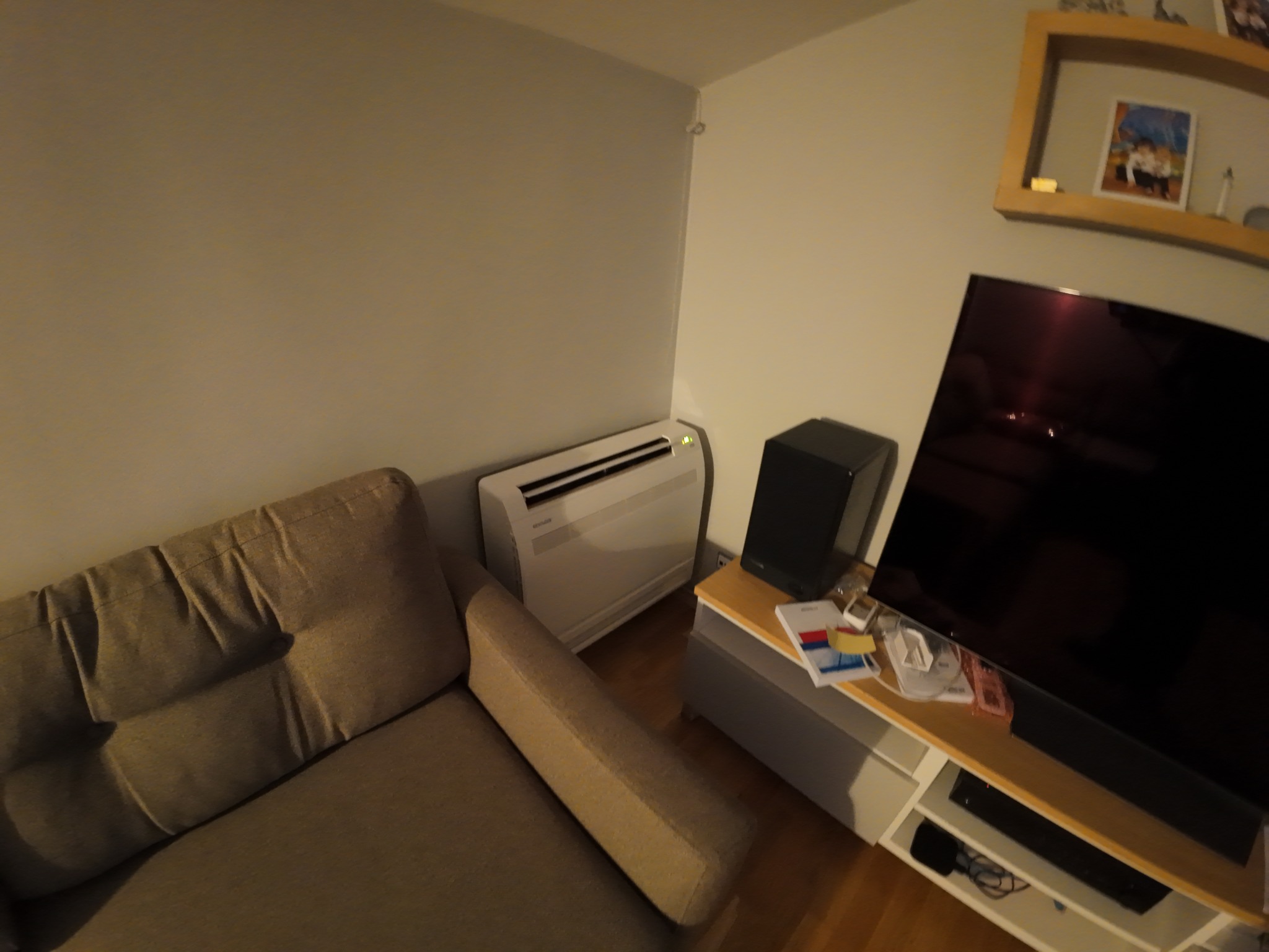 Biała jednostka klimatyzacji przypodłogowej w salonie, obok sofy i szafki RTV. Widok z szerokim kątem, ukazujący urządzenie w kontekście domowego wnętrza.