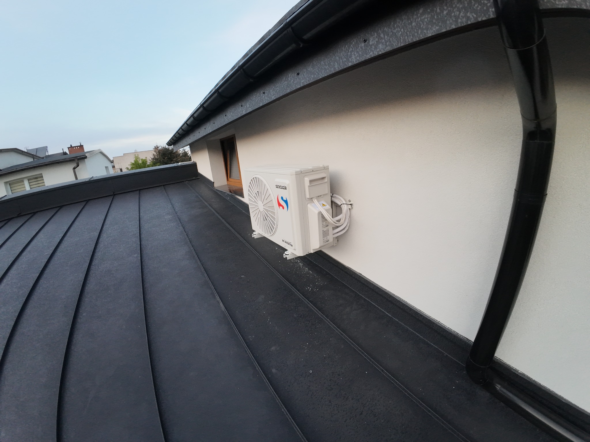 Biała jednostka zewnętrzna klimatyzacji Sinclair zamontowana na elewacji domu z czarnym dachem. Widok z dołu, z perspektywy dachu. Kabel zasilający schludnie ułożony.
