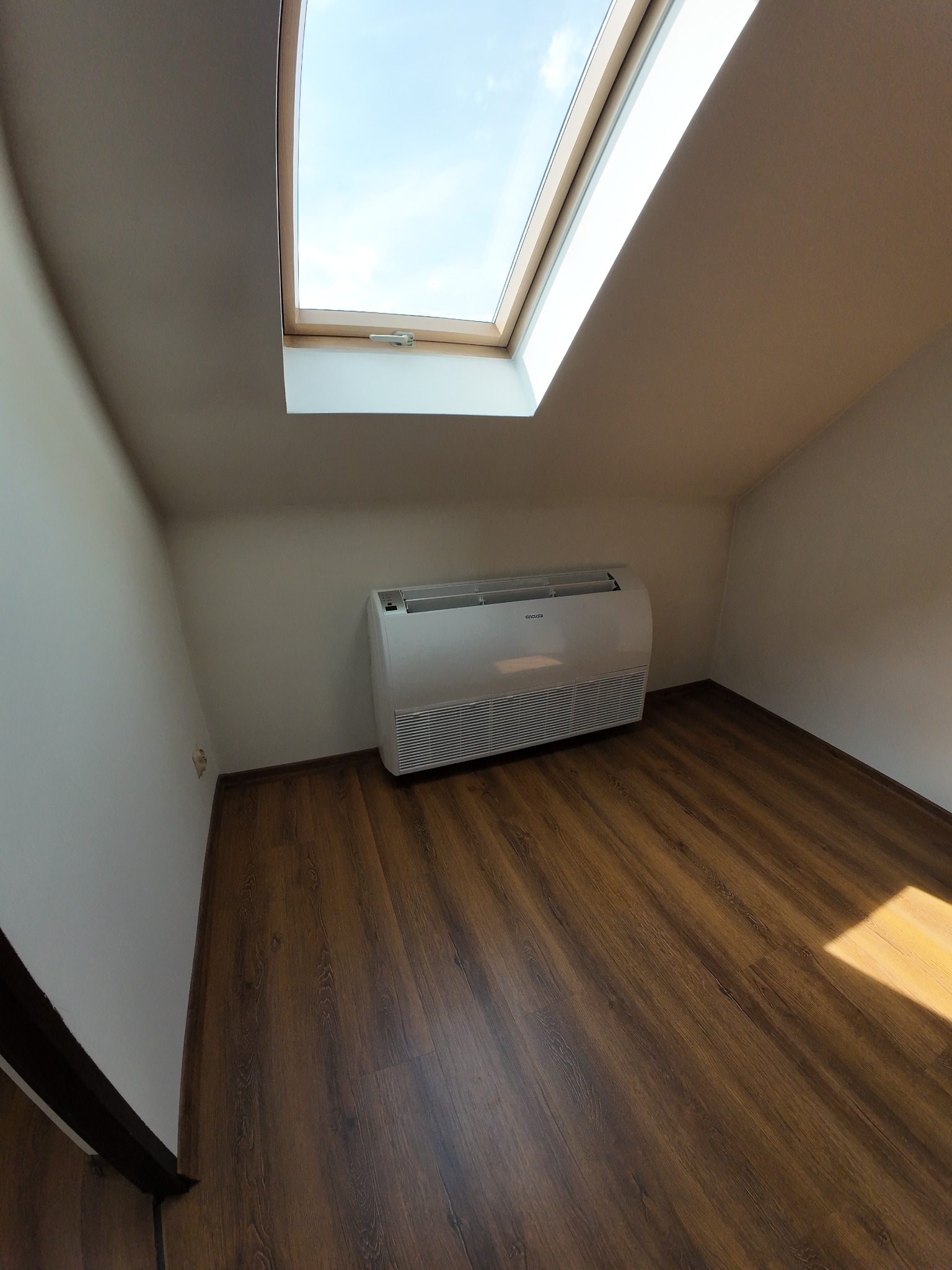 Biały klimatyzator Sinclair w jasnym pokoju na poddaszu z drewnianą podłogą i oknem dachowym. Widok szerokokątny.