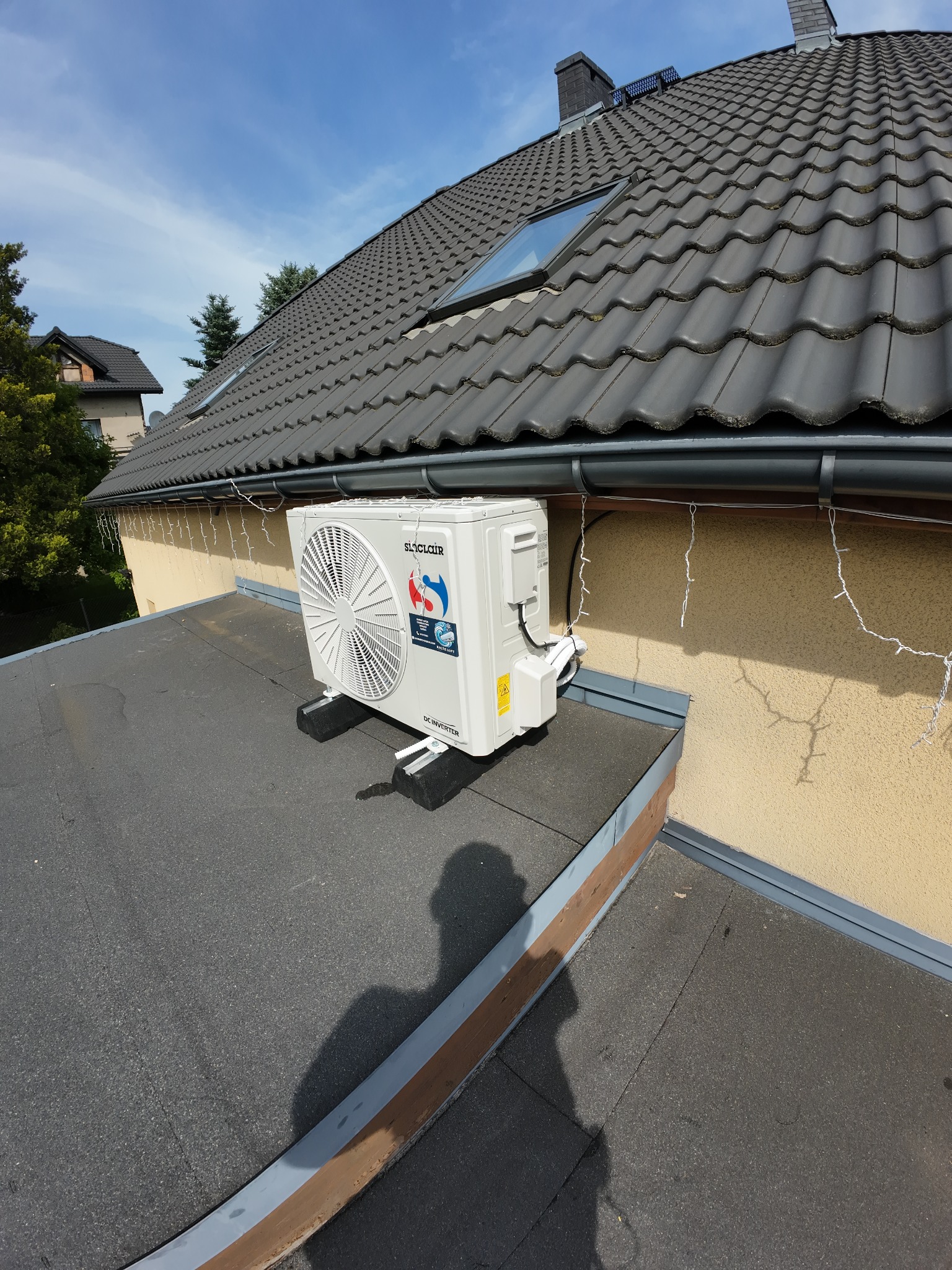 Biała jednostka zewnętrzna klimatyzacji Sinclair zamontowana na płaskim dachu budynku z szarą dachówką. Widoczne elementy instalacji i cień osoby robiącej zdjęcie.