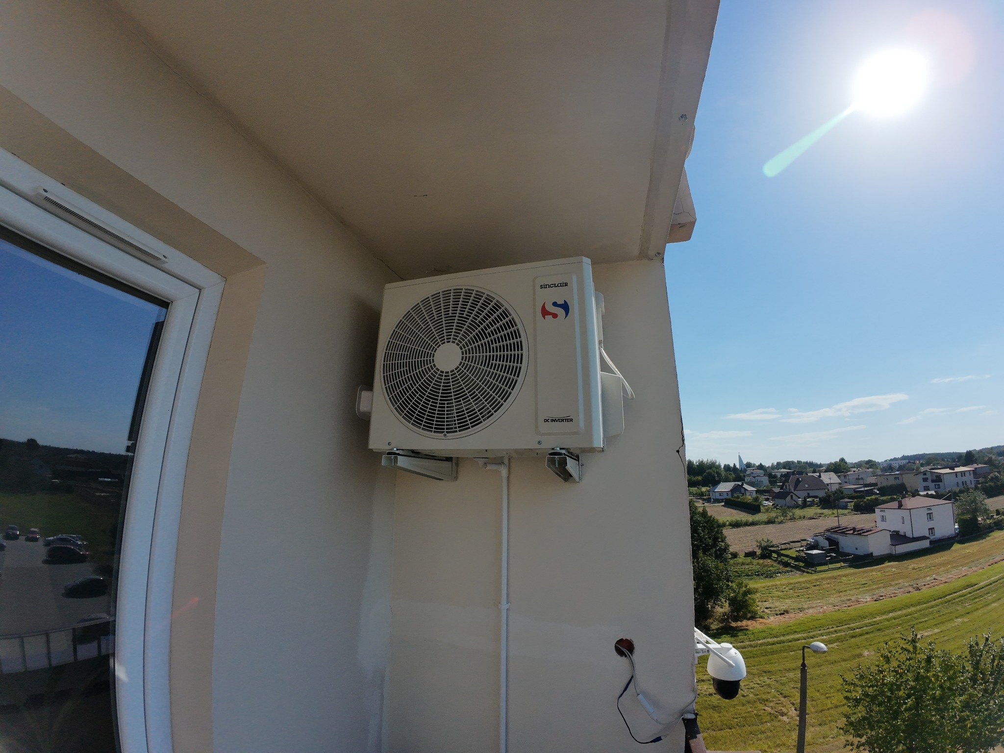 Zamontowana jednostka zewnętrzna klimatyzacji Sinclair na elewacji budynku, widok z boku, z widocznymi przewodami i elementami montażowymi. W tle krajobraz miejski i błękitne niebo.