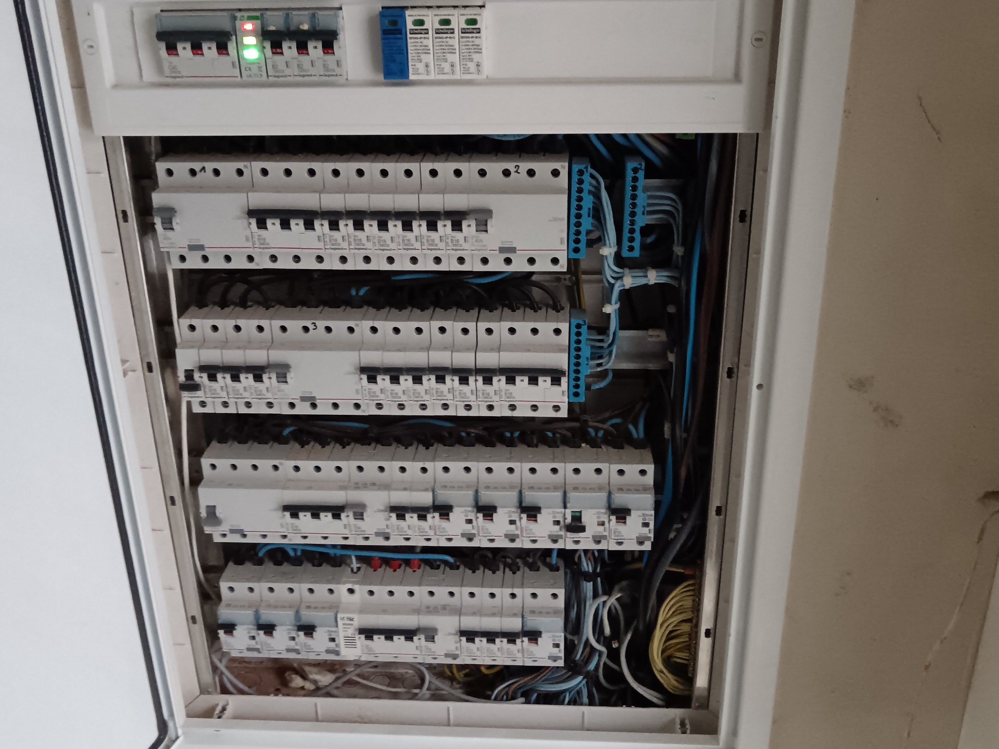 Rozdzielnica elektryczna z bezpiecznikami, widoczne przewody w kolorze niebieskim i żółtym. Górny rząd z diodami sygnalizacyjnymi. Biała obudowa.