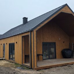 Oleksandr Matvieienko - Surowy, drewniany dom z ciemnym dachem na działce. Widoczne elementy konstrukcyjne i otoczenie w trakcie prac wykończeniowych. Ujęcie z zewnątrz, w pochmurny dzień.