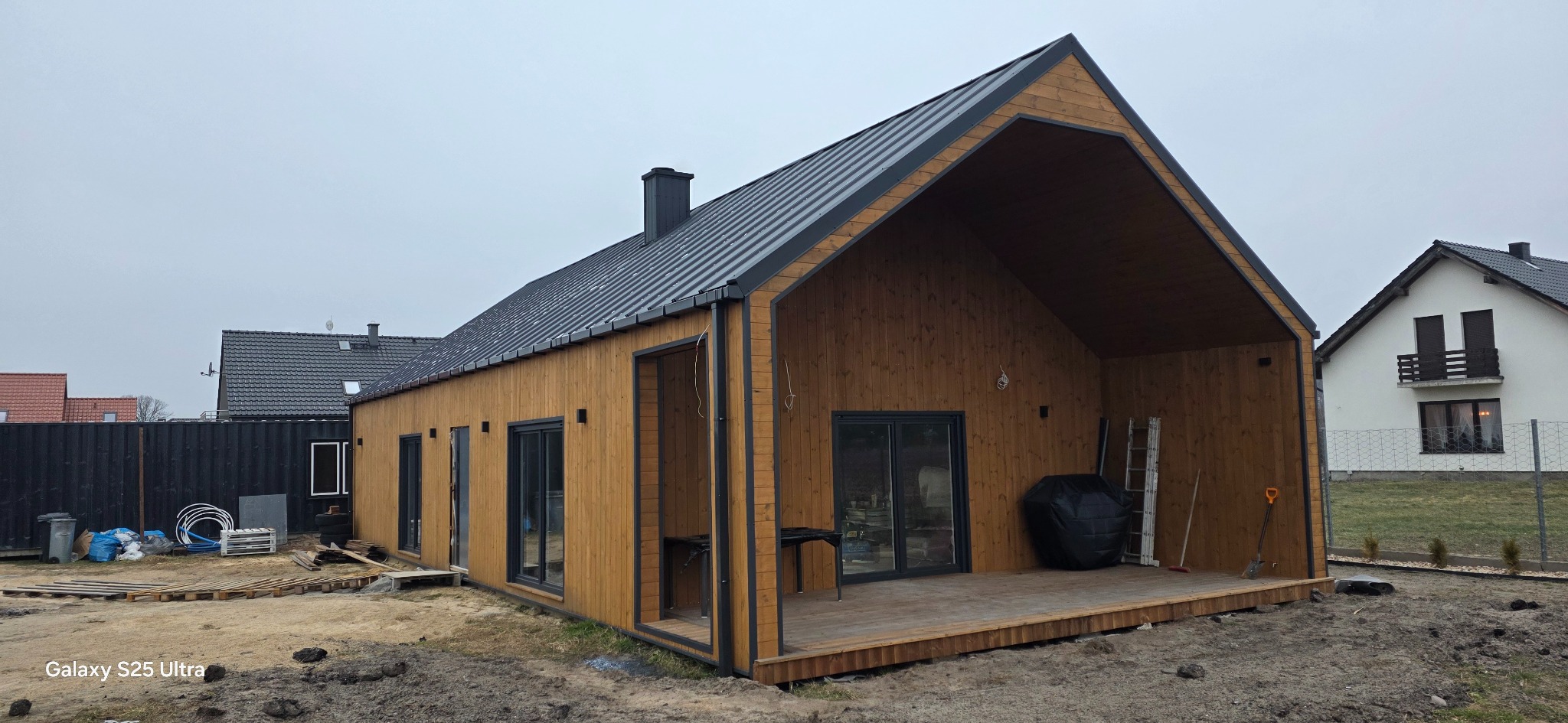 Surowy, drewniany dom z ciemnym dachem na działce. Widoczne elementy konstrukcyjne i otoczenie w trakcie prac wykończeniowych. Ujęcie z zewnątrz, w pochmurny dzień.