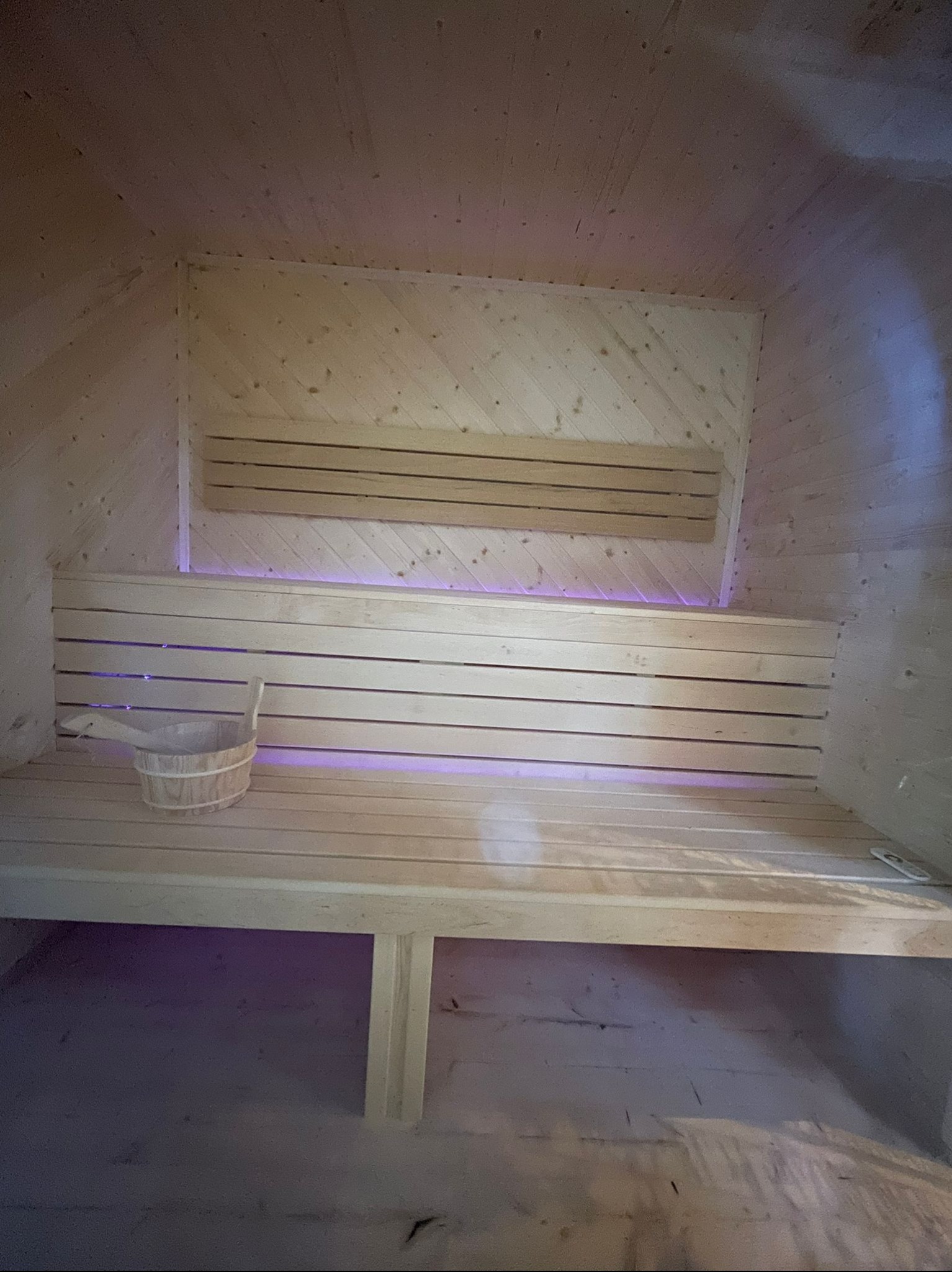 Wnętrze sauny z drewnianym wykończeniem, ławkami i wiadrem z chochlą. Subtelne, fioletowe podświetlenie dodaje klimatu. Widok z perspektywy osoby stojącej w saunie.