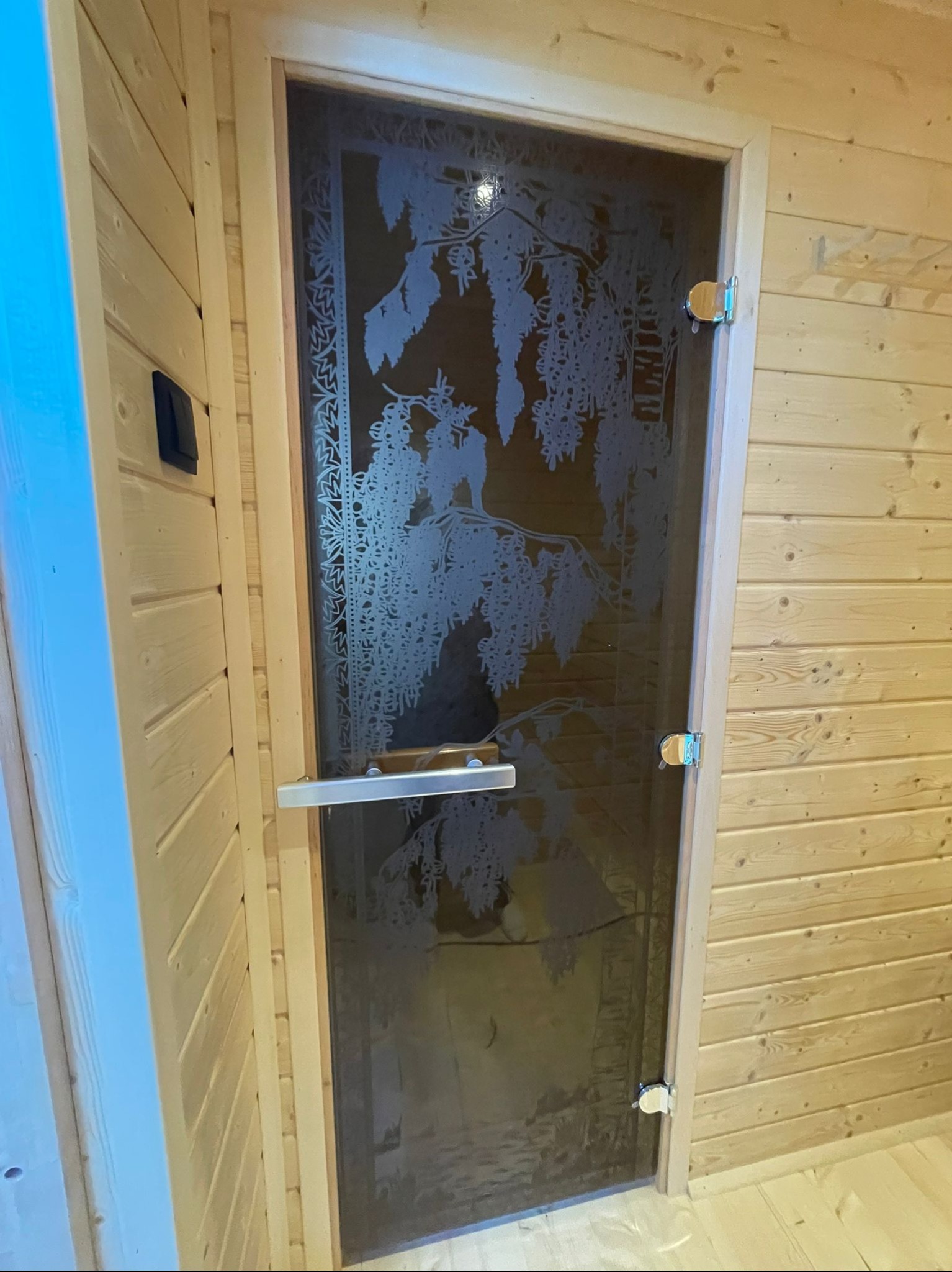 Szklane drzwi do sauny z dekoracyjnym motywem roślinnym, zamontowane w drewnianej konstrukcji z jasnych desek. Widoczne metalowe okucia i uchwyt.