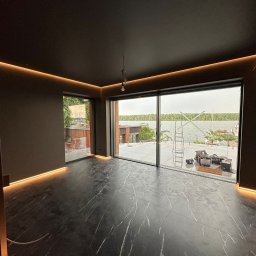 Usługi Elektryczne Dariusz Malenga - Nowoczesne wnętrze z czarną marmurową podłogą i subtelnym oświetleniem LED wzdłuż sufitu i podłogi. Widok na jezioro przez duże okno.